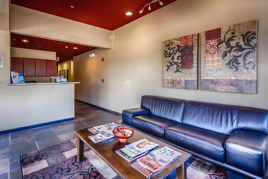 Modern dental waiting area with black leather sofa, red accent ceiling, artwork, and magazine table