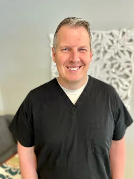 Professional portrait of a smiling male dental provider in black scrubs against a light patterned background