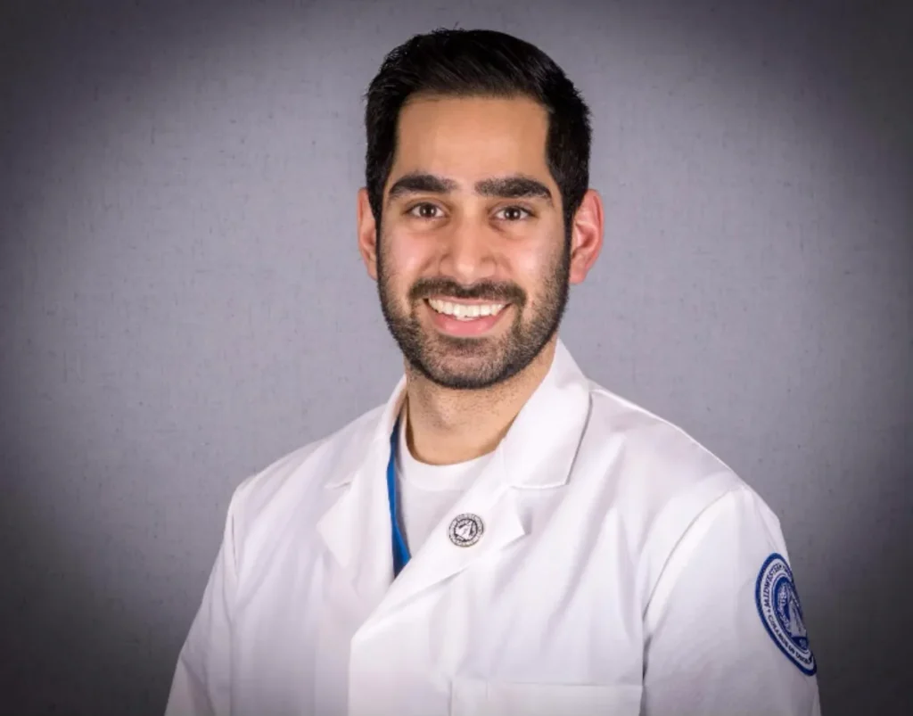 Professional headshot of a male dental team member in a white clinical coat against a gray studio background