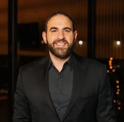 Professional headshot of a smiling male dental team member in a dark button-down shirt against a warm background