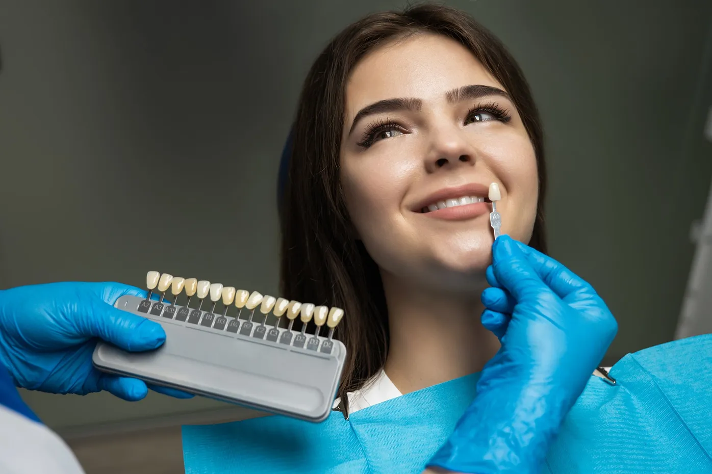 Young woman smiling during a dental shade matching or cosmetic consultation with a tooth sample guide nearby